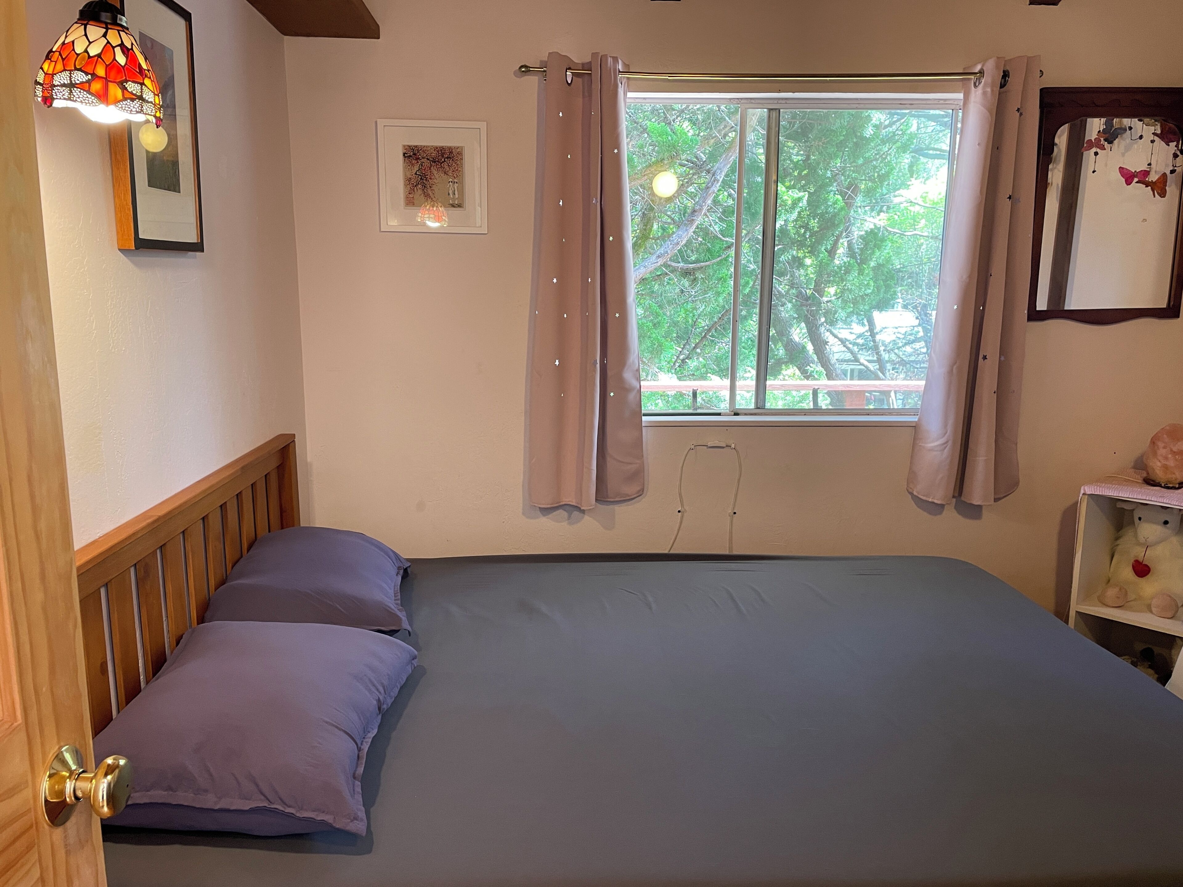 Un dormitorio con una cama, cabecera de madera, dos almohadas, una ventana con cortinas, un cuadro colgado en la pared y una lámpara fijada a la pared.