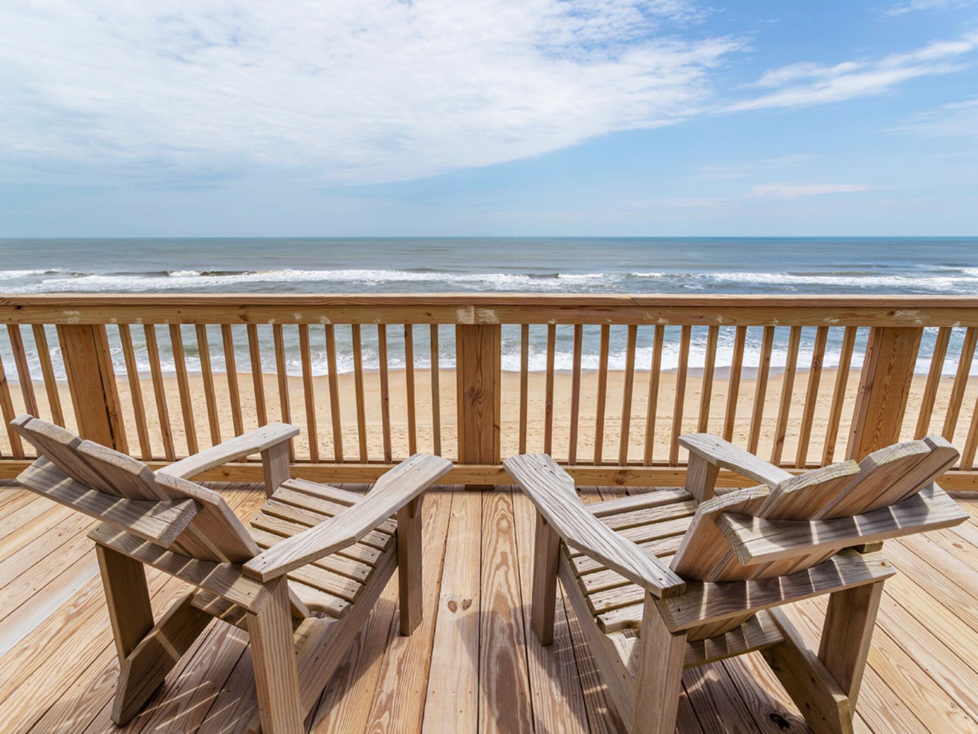 A wooden deck with two Adirondack chairs facing a beach with ocean waves.