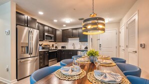 A modern kitchen with dark cabinets, stainless steel appliances, and a wooden dining table set with blue chairs and placemats. - VCR1-406 - 3BR Condo near all major attractions (Orlando)