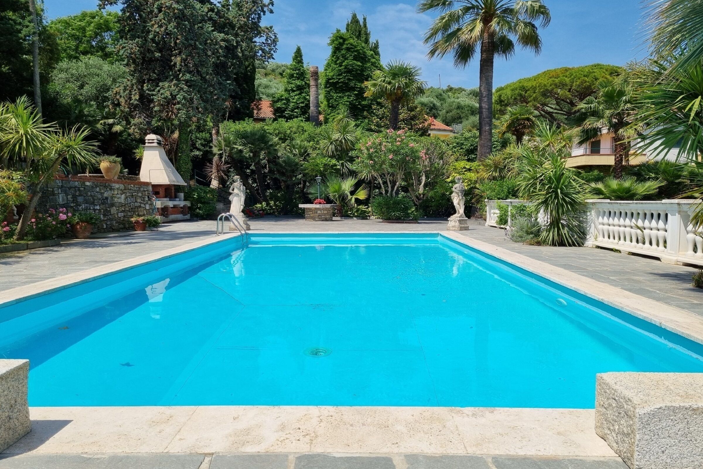 Ein rechteckiger Swimmingpool, umgeben von Steinmauern und üppigem Grün, mit einer Steinstruktur und Statuen im Hintergrund.