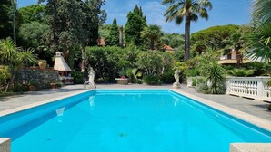 Una piscina rectangular rodeada de muros de piedra y exuberante vegetación, con una estructura de piedra y estatuas al fondo.