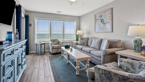 A living room with a sofa, armchairs, a coffee table, and a TV. There is a window with blinds, a ceiling fan, and a painting on the wall. - Luxurious & spacious family oceanfront condo w/great views, pools, in popular Cherry Grove (North Myrtle Beach)