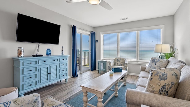 A living room with a blue cabinet, a flat-screen TV, a ceiling fan, a window with blinds, and a sofa with patterned cushions. - Luxurious & spacious family oceanfront condo w/great views, pools, in popular Cherry Grove (North Myrtle Beach)