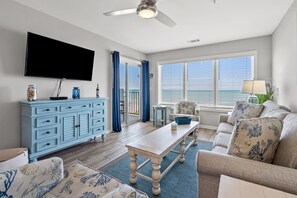 A living room with a blue cabinet, a flat-screen TV, a ceiling fan, a window with blinds, and a sofa with patterned cushions.