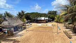 Ein Strandbereich mit einem Sandvolleyballfeld, strohgedeckten Gebäuden und Palmen.