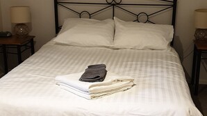 Una cama bien hecha con sábanas blancas, una toalla doblada y un albornoz negro sobre la cama. Encima de la cama, un cuadro que muestra a dos personas caminando por una calle.