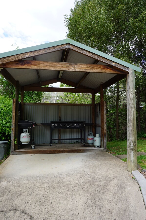 A covered outdoor barbecue area with a grill, propane tanks, and a concrete floor.