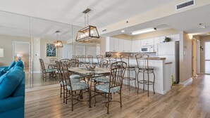 Un vaste espace de salle à manger avec une grande table en bois et des chaises assorties, une cuisine dotée de meubles blancs et un luminaire au plafond qui apporte beaucoup de lumière.