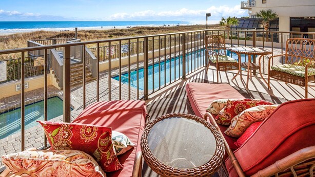 A balcony with a pool, wicker furniture, and a view of the beach and ocean.