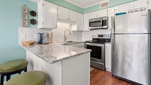A kitchen with white cabinets, a stainless steel refrigerator, a microwave, and an oven. There are two bar stools and a coffee maker on the counter.
