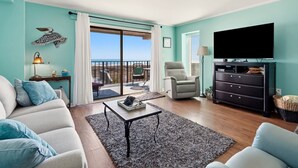 A living room with a sofa, a chair, a coffee table, a TV, and a view of the beach through a sliding door.