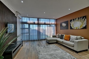 A modern living room with a sectional sofa, a flat-screen TV, and large windows with sheer curtains.