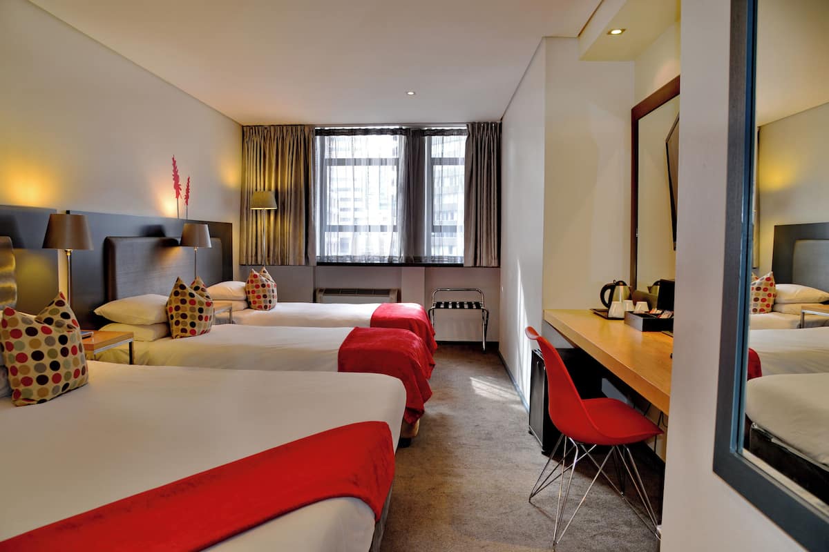 A hotel room with three beds, a desk, a chair, and a large window with curtains.