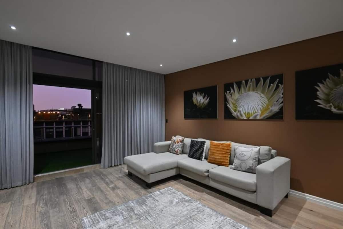 A modern living room with a sectional sofa, a large window with curtains, and three wall-mounted flower paintings.