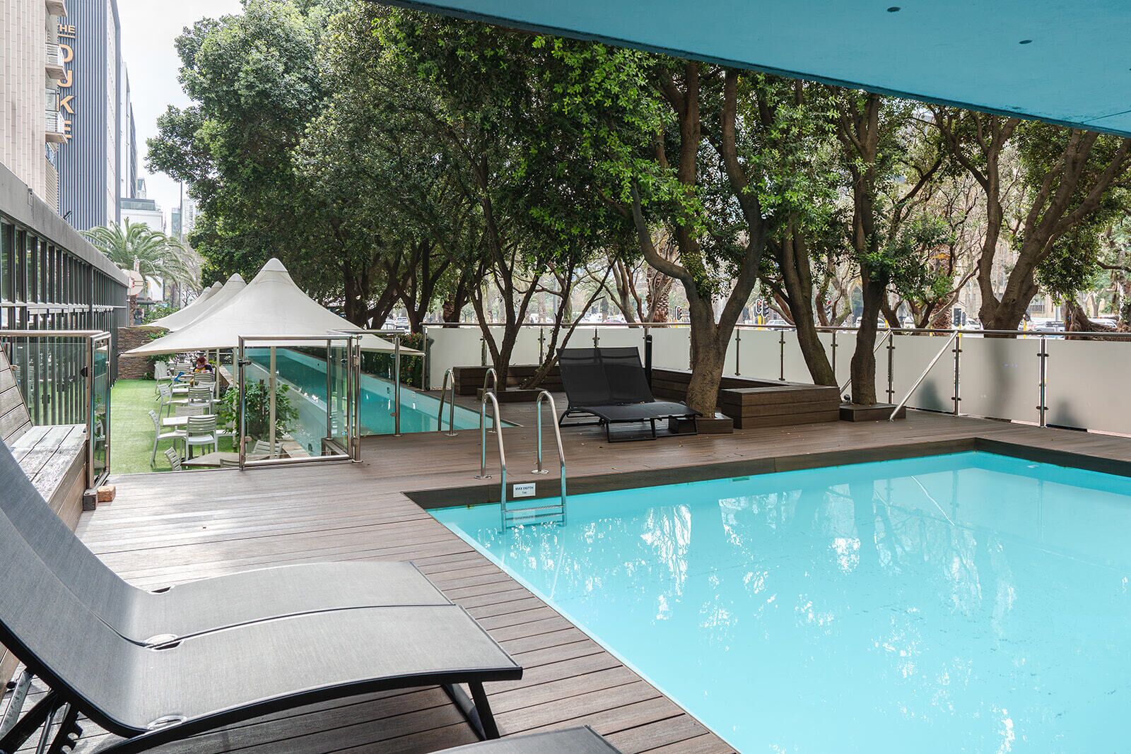 Una piscina con sillas de descanso y un cerramiento de vidrio, rodeada de árboles y un edificio.