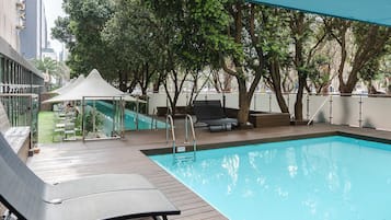 Una piscina con sillas de descanso y un cerramiento de vidrio, rodeada de árboles y un edificio.