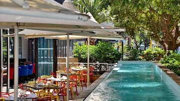 Zona de piscina al aire libre con mesas y sillas, rodeada de vegetación y un edificio con grandes ventanales.