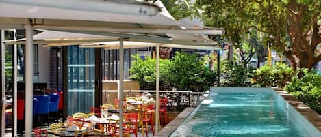 Zona de piscina al aire libre con mesas y sillas, rodeada de vegetación y un edificio con grandes ventanales.