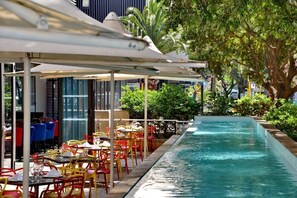 An outdoor pool area with tables and chairs, surrounded by greenery and a building with large windows. - Fountains Hotel by Neighbourgood (Cape Town)