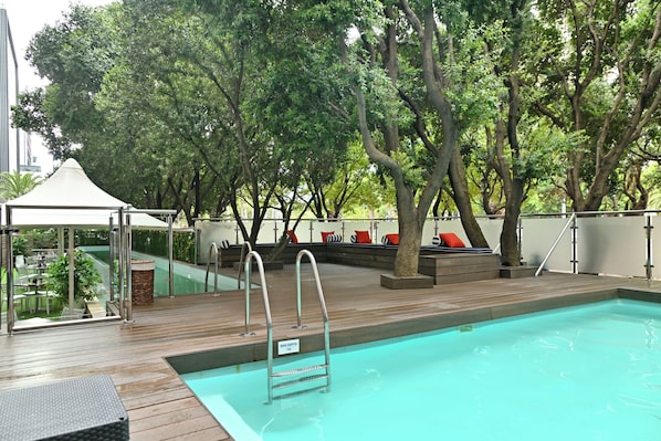 Piscina no terraço com deck de madeira, área de estar coberta e estrutura de vidro.