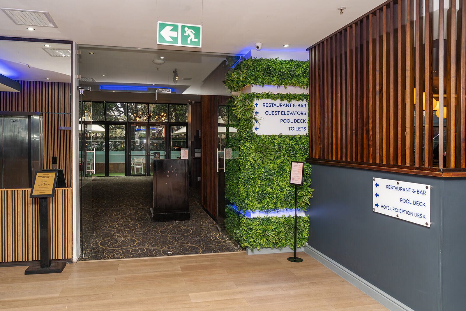 A hotel reception area with a wooden reception desk, a sign indicating the direction to the restaurant, bar, guest elevators, pool deck, and toilets.