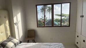 Un dormitorio con cama, una silla, un armario y ventana con vista a palmeras y una casa.