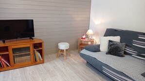Ett modernt vardagsrum med en platt-TV på ett träställ, en liten vit pall, ett sidobord med en lampa och en kattfigur, och en grå soffa med en randig pläd.