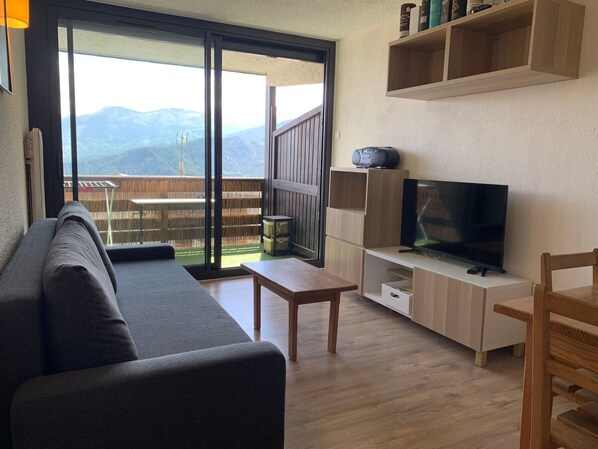 A living room with a sofa, a TV, a wooden table, and a balcony with a view of mountains.