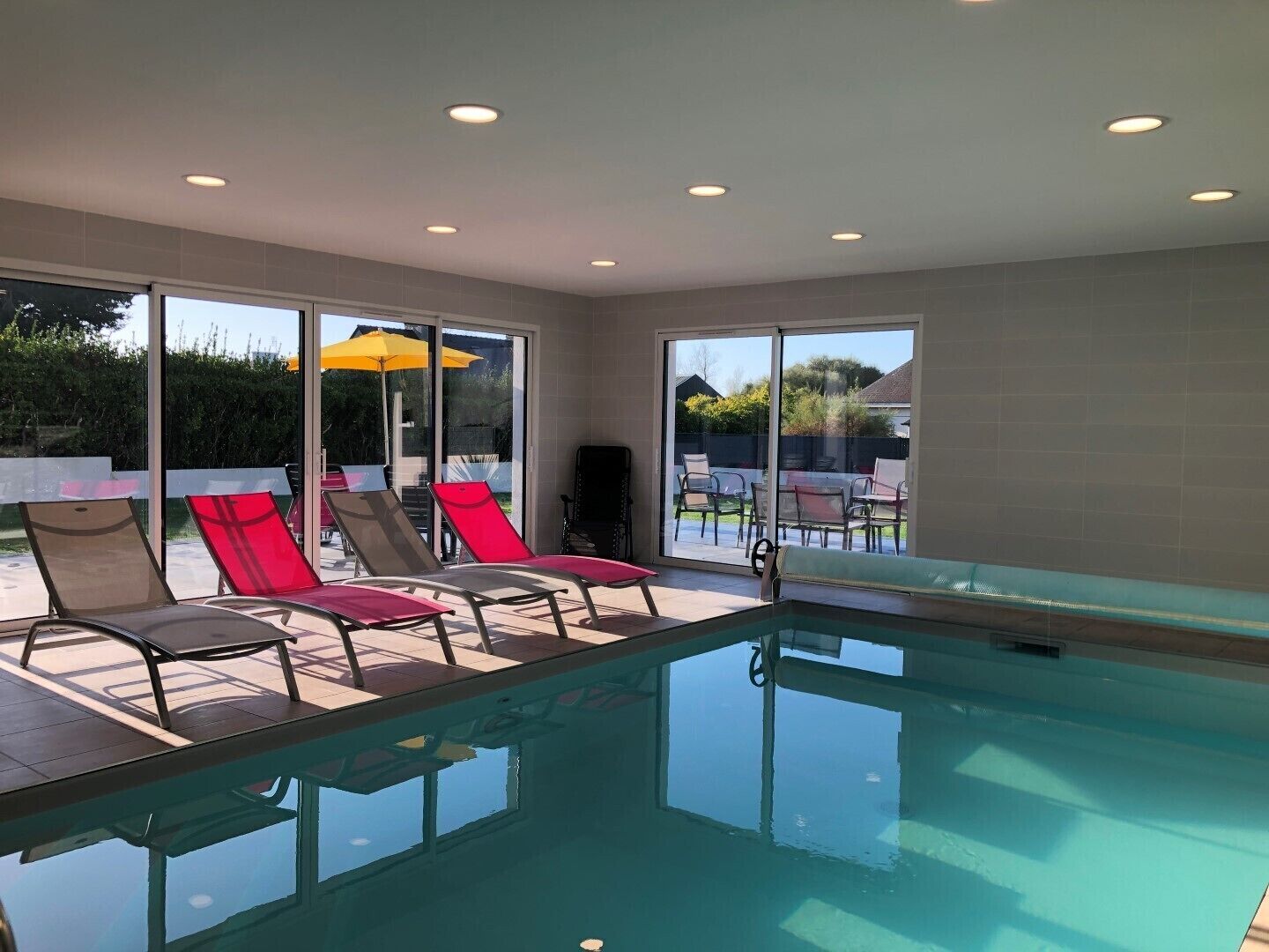 Una piscina interna con pareti in vetro trasparente, circondata da lettini e un ombrellone giallo.