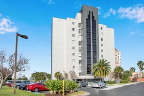 Un edificio moderno de varios pisos con una fachada central de vidrio vertical, rodeado de coches aparcados y palmeras.