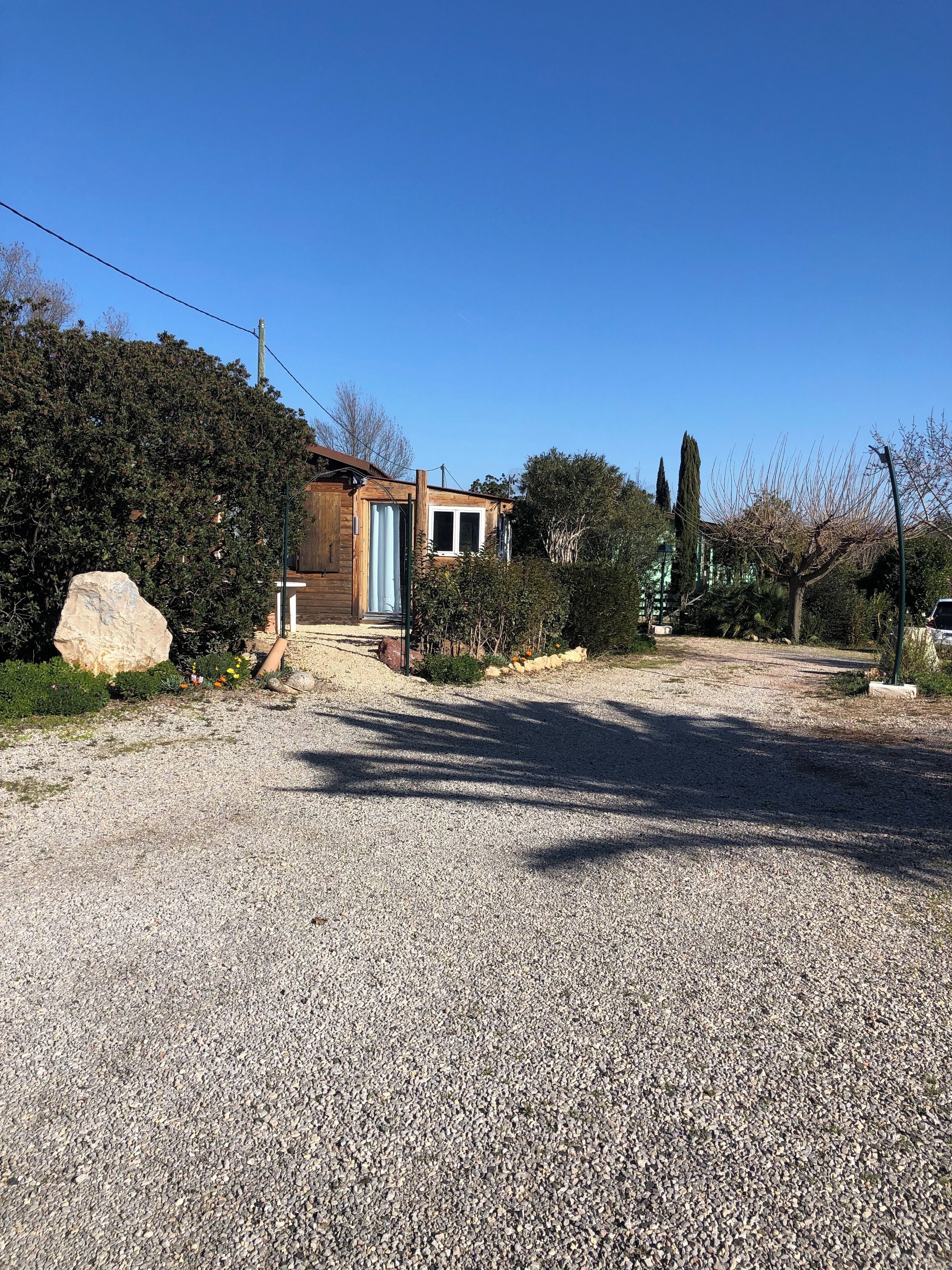 Un carril de grava que condueix a una casa de fusta amb una porta blava, envoltada de malesa abundant i una gran roca.