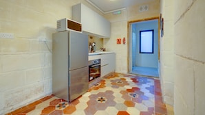 Een compacte keuken met een koelkast, oven en witte kasten. Een raam zorgt voor natuurlijk licht, en de vloer heeft een geometrisch tegelpatroon.