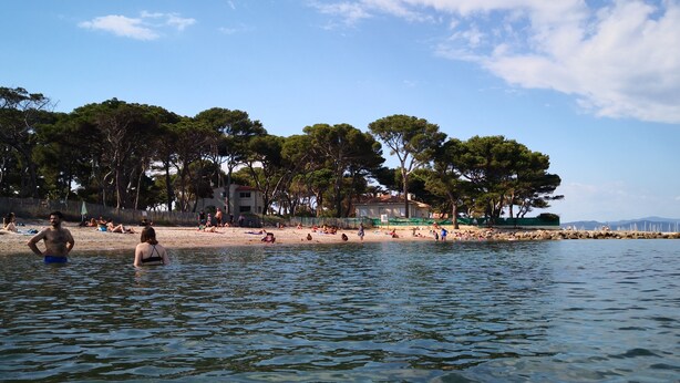 Una playa con gente tomando el sol y nadando, un edificio de techo rojo, y una hilera de árboles a lo largo de la orilla.