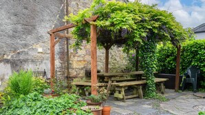 A stone building with a wooden pergola covered in green foliage and purple grapes, surrounded by potted plants and stone-paved patio with wooden picnic tables.