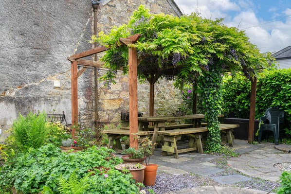A stone building with a wooden pergola covered in green foliage and purple grapes, surrounded by potted plants and stone-paved patio with wooden picnic tables.