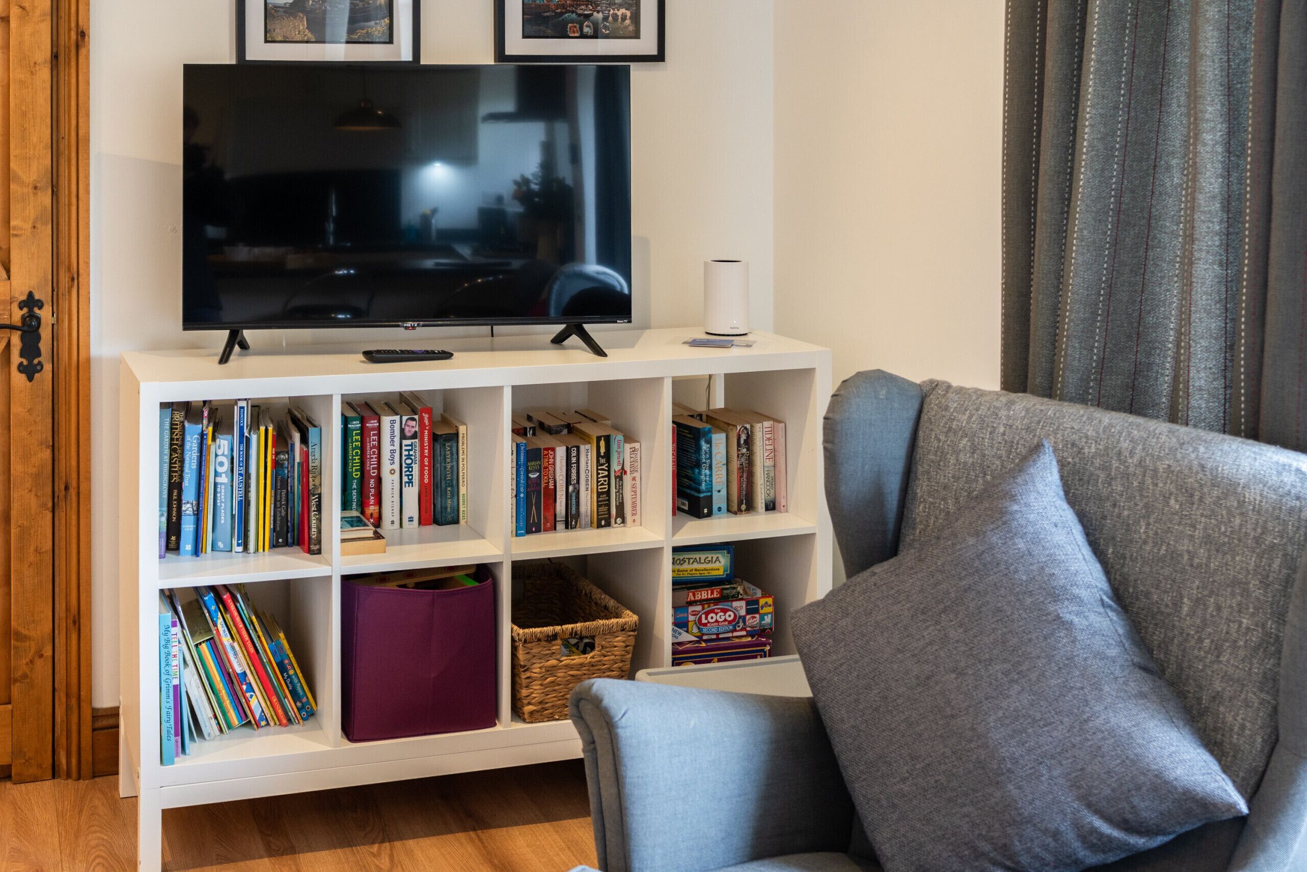 A living room with a flat-screen TV, a bookshelf, a grey armchair, and a wooden door.