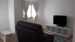Een woonkamer met een zwarte bank, een witte tv-meubel met een flatscreen televisie, een klein wit kastje en een raam met witte gordijnen.