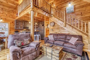 A wooden cabin interior with a sofa, a coffee table, a kitchen area, and a staircase.