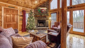 A cozy cabin interior with a fireplace, wooden walls, a Christmas tree, and a large window overlooking a deck.