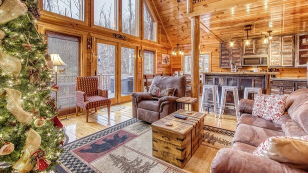 A cozy log cabin interior with a large wooden ceiling, a fireplace, a dining area, and a view of the surrounding forest.