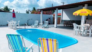 A swimming pool with two colorful lounge chairs, a red umbrella, and white plastic chairs and tables. - Casa de Praia Condomínio Fechado em Ipioca (Maceió)