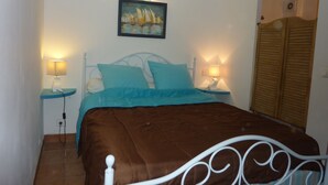 Een net opgemaakt bed met een bruine deken, twee blauwe kussens en een wit metalen hoofdbord. Er zijn twee nachtkastjes met lampen, en een schilderij van zeilboten aan de muur.