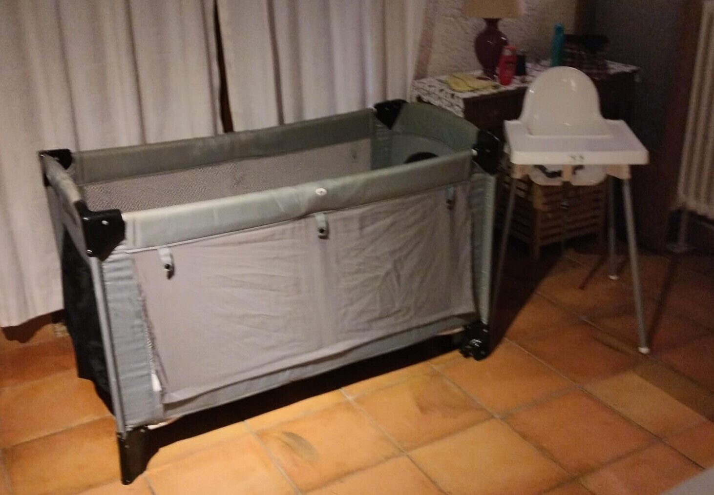 A baby changing station with a mesh top and a padded bottom, a white high chair, and a small table with a lamp and items on it.