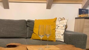 Una mesa de centro de madera con dos copas de vino, colocada frente a un sofá gris con un cojín amarillo.