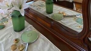 Une chambre avec un grand miroir, un lit recouvert de draps blancs, une table de chevet ornée d’un vase vert et d’une nappe à motifs floraux, et une fenêtre laissant entrer la lumière du jour.