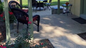 Un patio couvert aménagé avec du mobilier d’extérieur, une table ornée d’une citrouille, et une vue sur une clôture blanche et des bâtiments en arrière-plan.