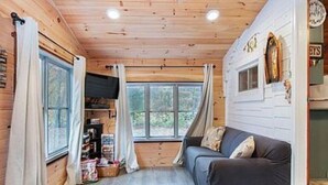 A cozy cabin interior with a wooden ceiling, a sofa, a window, and a bookshelf. - 1-bedroom cabin near Huron-Manistee National Forest (Newaygo)