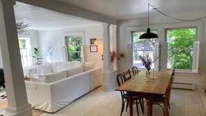 A spacious living room with a white sofa, a wooden dining table, and a large window providing a view of greenery. - Renovated Catskills Farmhouse - hiking, swimming, quaint towns, 1st floor living (Denver)