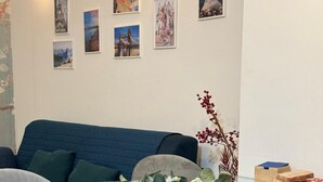 En moderne stue med et sofabord i glas, grå polstrede stole, en blå sofa og en væg pyntet med billeder i rammer.