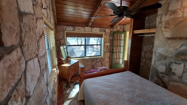 A rustic room with a wooden ceiling, stone walls, a bed, a dresser, and a window with a view of trees. - Cozy, private waterfront stone cabin on the north shore of Lake Travis (Lago Vista)